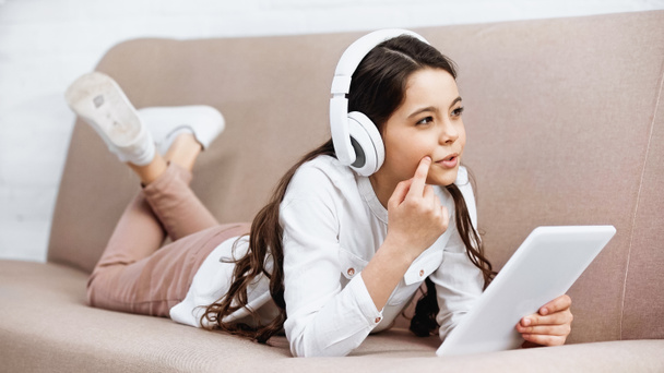 Pensive preteen girl in headphones holding digital tablet on couch at home  - Fotó, kép