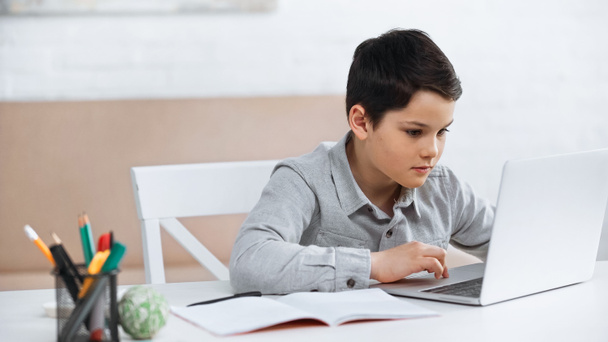 Poika käyttää kannettavaa tietokonetta lähellä muistikirjaa kotona  - Valokuva, kuva