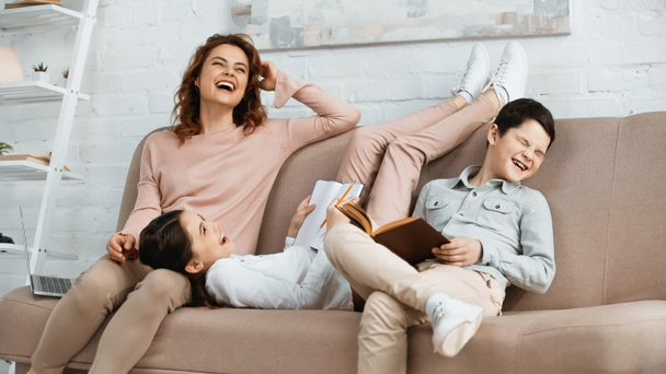 Positive woman sitting near kids with book, notebook and laptop on couch at home  - Photo, Image