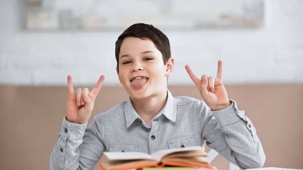 Preteen boy zeigt Rock-Geste und streckt die Zunge in der Nähe von Büchern auf verschwommenem Vordergrund  - Foto, Bild