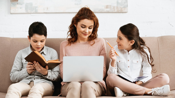 Vidám lány jegyzetfüzettel és ceruzával ül anya mellett laptoppal és testvér olvasókönyv  - Fotó, kép