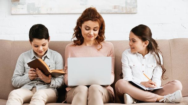Smiling kids doing homework near mother with laptop on couch  - Photo, Image