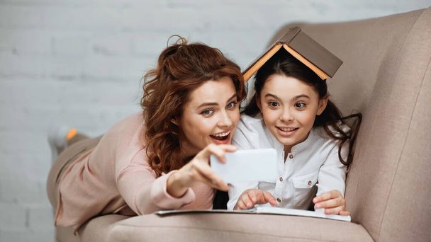 Anya és lánya szelfit készít okostelefonon könyvvel és jegyzetfüzettel a kanapén  - Fotó, kép