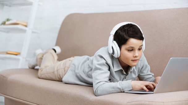 Niño preadolescente en auriculares con ordenador portátil en el sofá   - Foto, Imagen