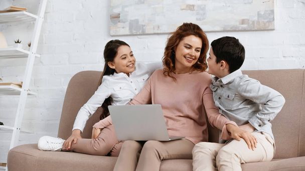 Smiling mother with laptop hugging kids on couch  - Fotografie, Obrázek