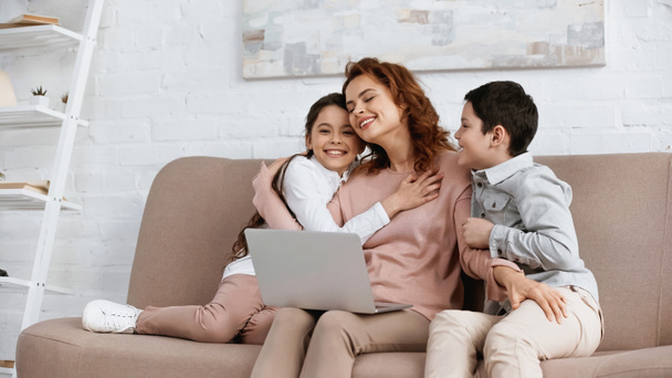 Woman hugging smiling children near laptop on couch  - 写真・画像