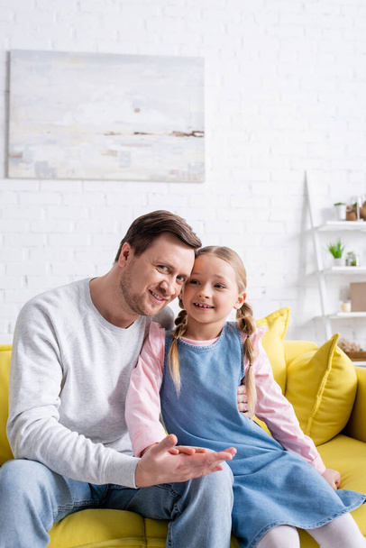 happy child touching hand of father hugging her on sofa at home - Foto, Bild