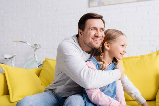 joyful man looking away while hugging daughter on couch at home - Foto, imagen