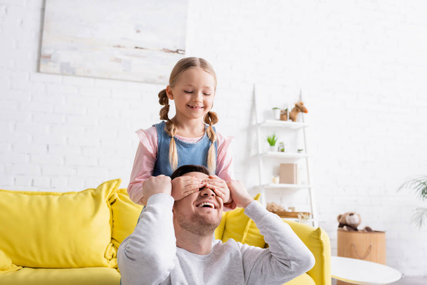 lachend meisje dekking ogen van lachende vader terwijl plezier hebben thuis - Foto, afbeelding