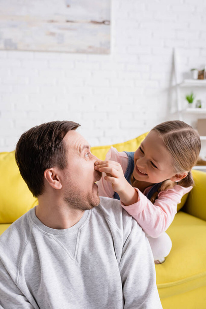 chica juguetona taponamiento nariz de padre mientras se divierten en la sala de estar - Foto, Imagen