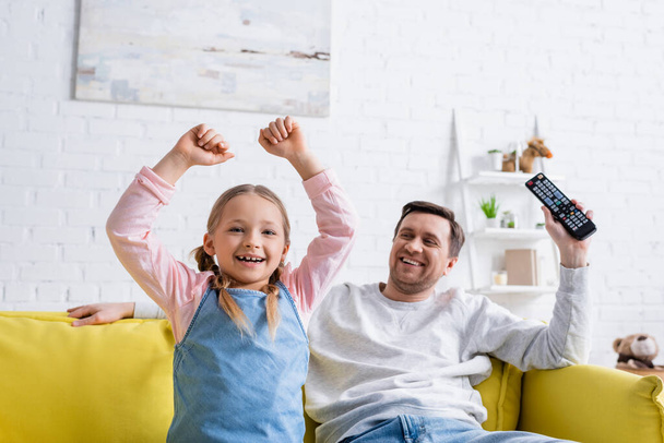 cheerful girl showing triumph gesture while watching tv near dad at home - Φωτογραφία, εικόνα