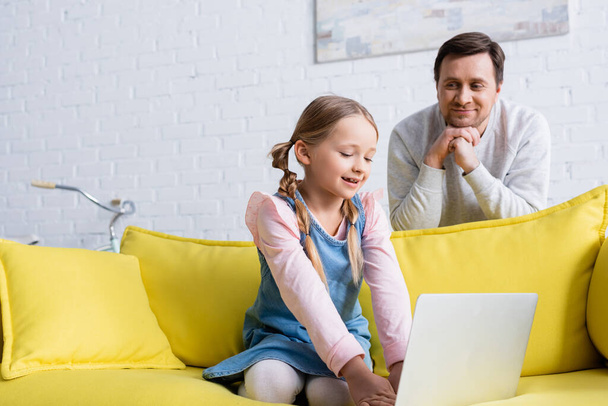joyful man looking at daughter using laptop on sofa - Photo, Image