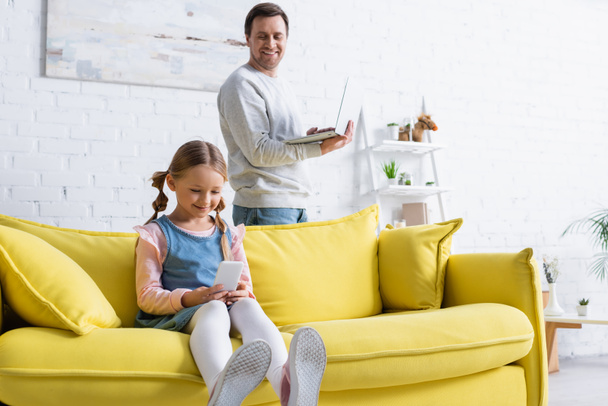 smiling man with laptop looking at daughter using mobile phone on couch - Fotó, kép