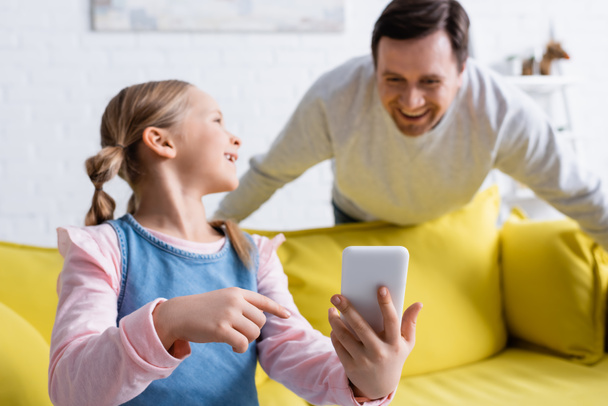 smiling girl pointing at mobile phone near father laughing on blurred background - Photo, image