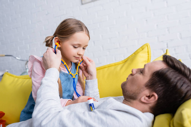 muž ležící na pohovce, zatímco dcera zkoumá ho s hračkou stetoskop - Fotografie, Obrázek