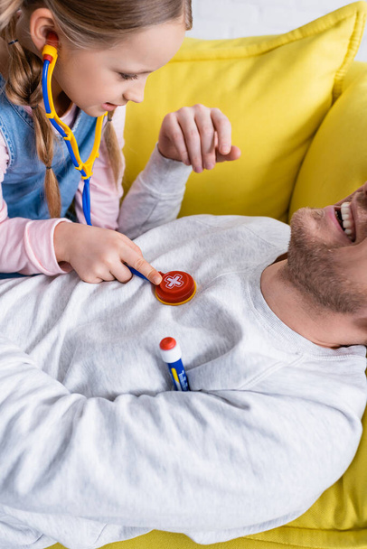uomo ridente sdraiato sul divano vicino figlia esaminandolo con stetoscopio giocattolo - Foto, immagini