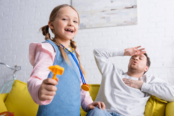 excited girl holding toy reflex hammer while playing doctor near dad pretending sick on blurred background - Photo, Image