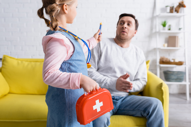 girl holding toy syringe and first aid kit near father pretending scared on blurred background - Photo, Image