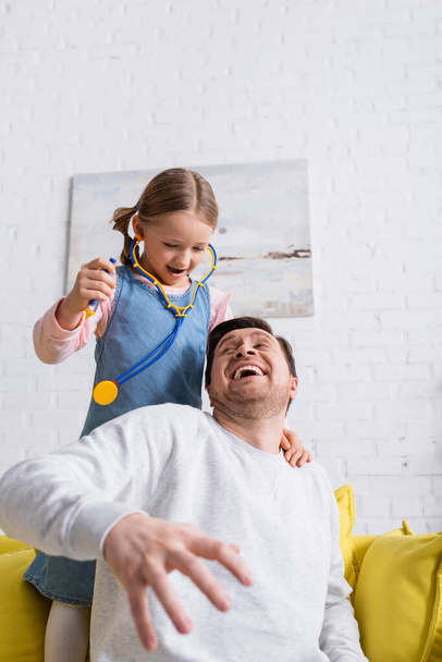father pretending scared near daughter holding toy syringe while playing doctor, blurred foreground - Фото, изображение