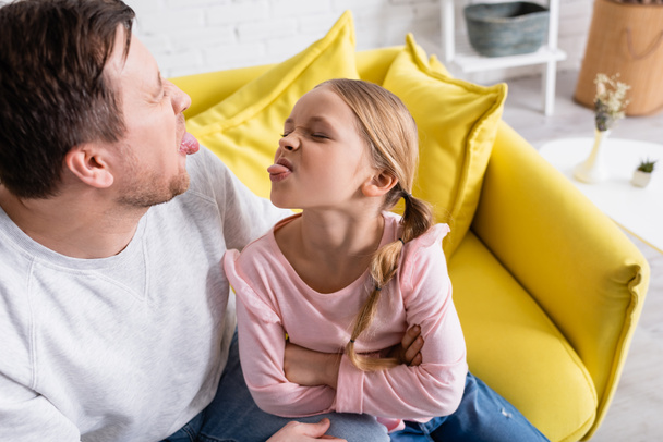 father and daughter sticking out tongues to each other at home - Фото, изображение