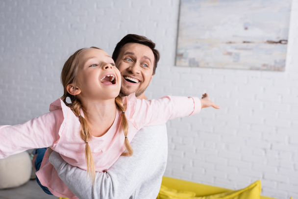 ευτυχισμένος άνθρωπος κρατώντας έκπληκτος κόρη δείχνει μύγα χειρονομία, ενώ έχει τη διασκέδαση στο σπίτι - Φωτογραφία, εικόνα