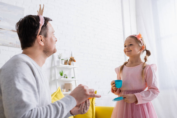 vrolijk meisje in speelgoed kroon kijken naar vader op wazig voorgrond - Foto, afbeelding