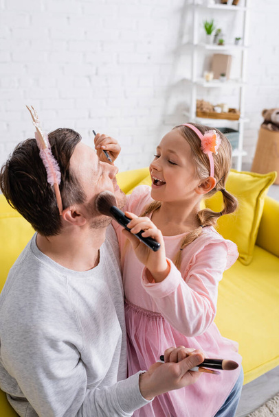cheerful daughter applying makeup to father wearing toy crown - Fotografie, Obrázek