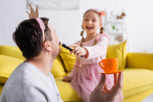 ενθουσιασμένοι κορίτσι κάνει μακιγιάζ στον πατέρα φορώντας στέμμα παιχνίδι, θολή φόντο - Φωτογραφία, εικόνα