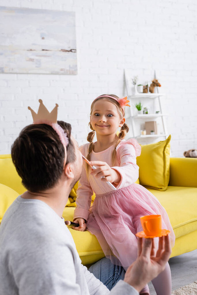 схвильована дівчина робить макіяж з батьком, тримаючи іграшкову чашу на розмитому передньому плані. - Фото, зображення