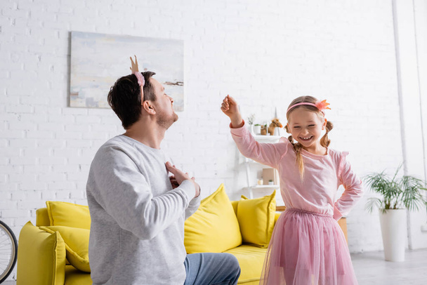 man in toy crown holding hands on chest while inviting cheerful daughter to dance - Фото, зображення