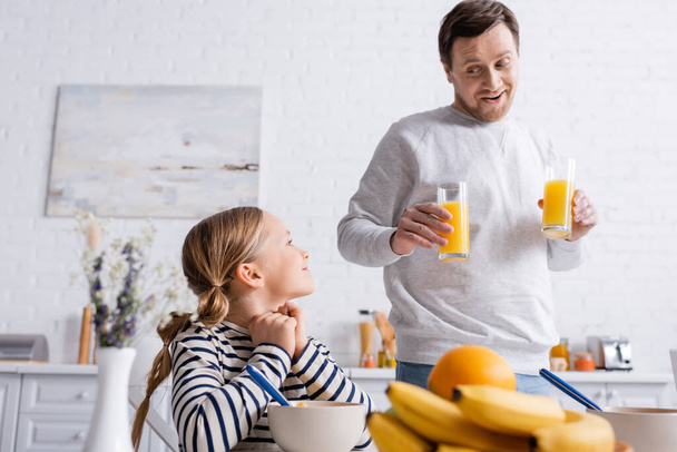 cheerful father holding orange juice near daughter and fruits on blurred foreground - Fotoğraf, Görsel
