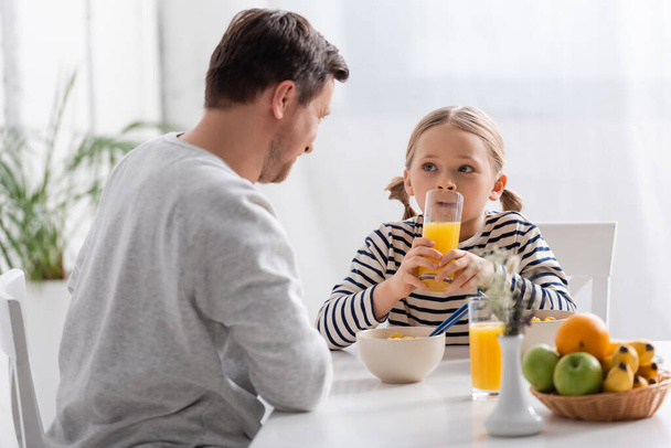 girl drinking orange juice near father and fruits on blurred foreground - Photo, Image