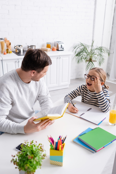 man holding book near excited daughter doing homework in kitchen - Photo, image