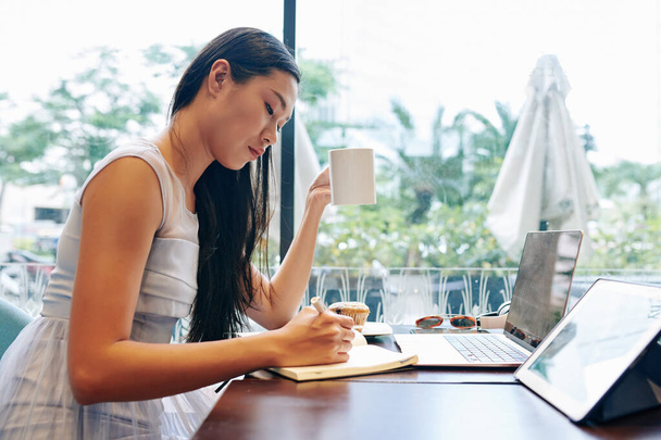 Серьезная молодая женщина сидит за журнальным столиком, пьет кофе и пишет планы на день в блокноте - Фото, изображение