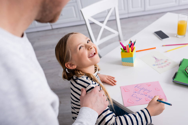 father touching shoulder of cheerful girl drawing happy womens day card, blurred foreground - Фото, изображение
