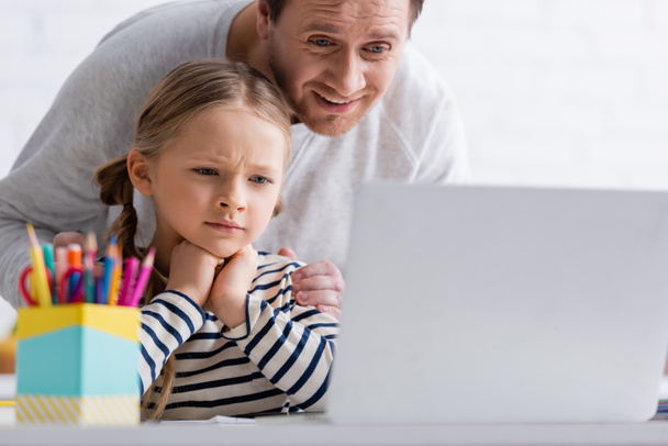 smiling man touching thoughtful daughter during online lesson at laptop on blurred foreground - Photo, Image