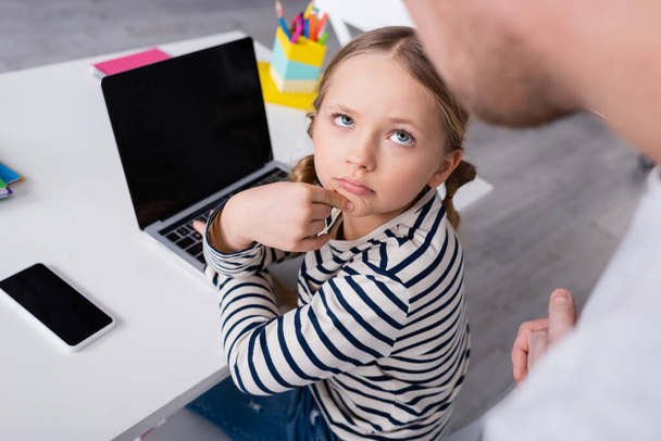 schoolgirl looking at father on blurred foreground while sitting near laptop - Photo, Image