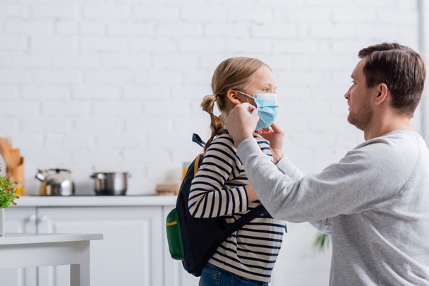 man putting on protective mask on daughter with backpack - Фото, изображение