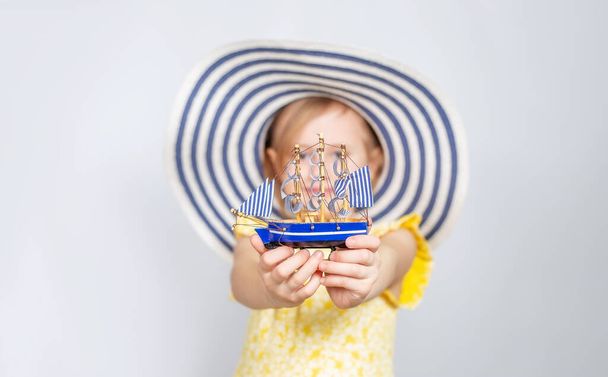 A Little Caucasian girl in a wide-brimmed hat holds out a toy boat forward. Focus on the foreground. The concept of summer vacation, travel, opening of the beach season.. - Fotografie, Obrázek