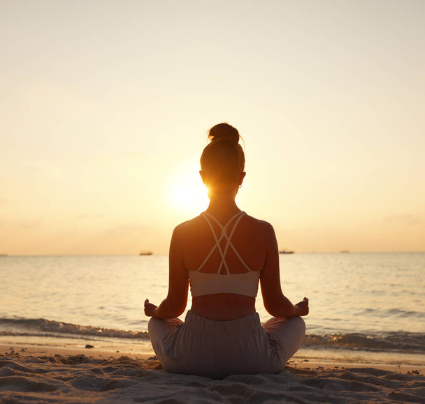 Погляд на самицю, що сидить у Падмасані під час медитації на узбережжі проти неба під сонцем. - Фото, зображення
