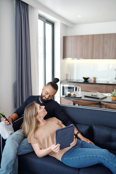 Szczęśliwa młoda mieszana para odpoczywająca na kanapie w domu i sprawdzająca media społecznościowe na smartfonie i tablecie - Zdjęcie, obraz