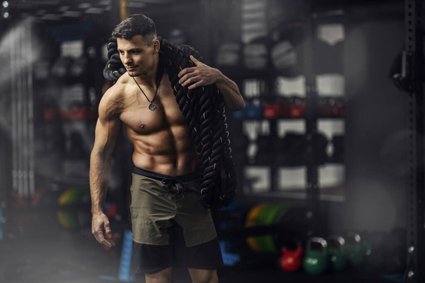 A close-up shot of a sexy man with a bare upper body on his shoulder carries a fitness rope in the dark atmosphere of a gym. Strong body shape and healthy sports life, fitness lover, trainer - Photo, Image