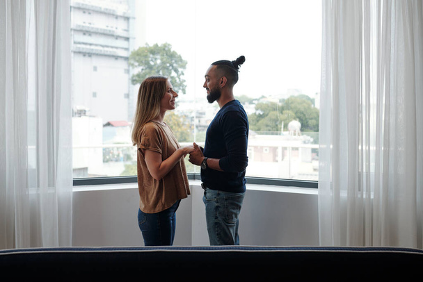 Счастливая влюбленная смешанная пара, стоящая на балконе, держась за руки и глядя друг на друга - Фото, изображение