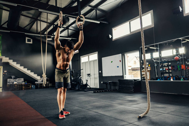Der muskulöse Körper eines Mannes ohne T-Shirt beim Training. Ein Fitness-Mann steht mit nacktem Oberkörper in der Mitte der Turnhalle und stützt seine Hände auf Reifen. Volle Länge der muskulösen Körper - Foto, Bild