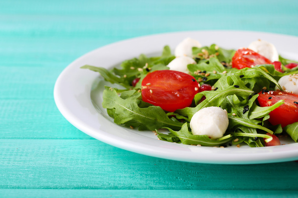 Green salad made with  arugula, tomatoes, cheese mozzarella balls and sesame  on plate, on color wooden background - Foto, Imagem