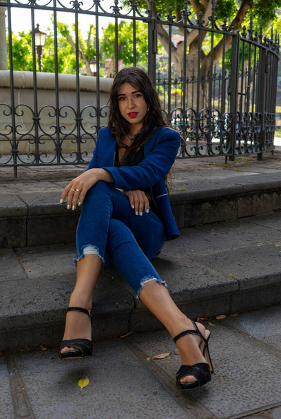 Young woman posing for a portrait sitting on the floor with her legs and arms crossed. She wears heels, jeans and an American jacket. In the background you can see a fountain from La Laguna, Teneri - 写真・画像