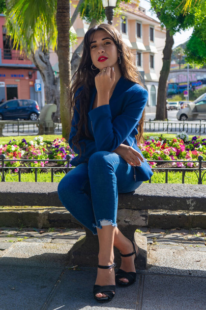 Young woman posing for a portrait. She wears an American jacket, jeans and heels. She is sitting on a bench in La Laguna, Tenerife. - Fotó, kép