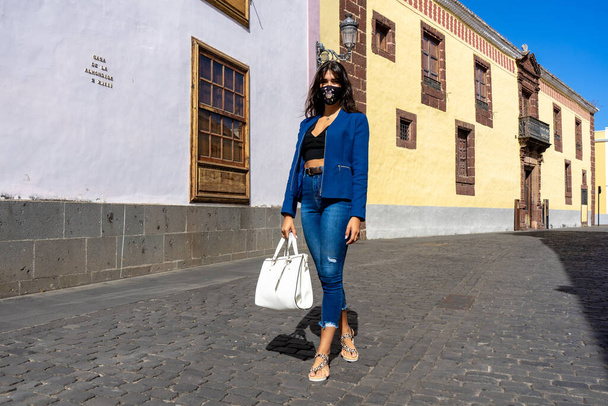 Young woman walking down a pedestrian street in La Laguna, Tenerife. She wears mask, jeans, an American jacket, a bag and sandals - Fotoğraf, Görsel