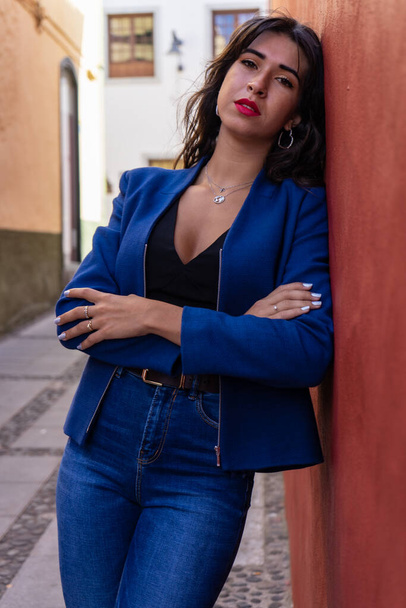 Young woman with arms crossed leaning against a red wall. She wears jeans and an American jacket. The photo was taken in La Laguna, Tenerife. - Foto, Imagem