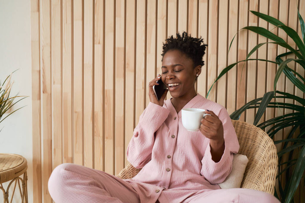 Happy young girl in the morning in cozy pink pajamas sits in a wicker chair with a cup of coffee and talks on the phone. - Foto, afbeelding